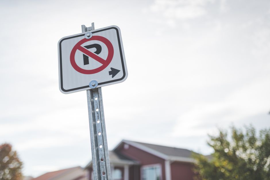 A metal no-parking sign installed on a pole outdoors, displaying a red circle with a diagonal line crossing out the letter P, indicating no parking, with a small black arrow pointing to the right. The sign is situated in a residential area, with a partly cloudy sky overhead, a house with a sloped roof, and some trees visible in the background. This image is relevant to home relocation and furniture transport, as it depicts an area where loading and unloading for removals services by Man with van Bedford Park may occur, highlighting the importance of observing parking rules during the moving process.