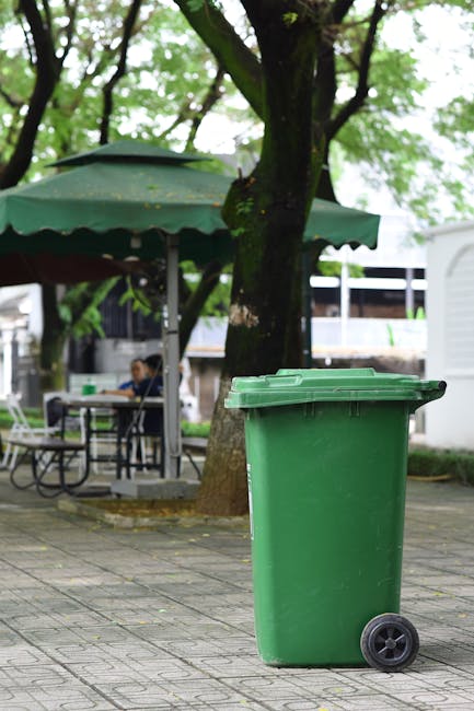 A green wheeled rubbish bin with a closed lid positioned on a paved outdoor area beneath a large tree with green leaves, with a park-like setting including outdoor seating with a round table, chairs, and a large umbrella, and a few people sitting in the background. The scene appears to be in an outdoor public space in Bedford Park, with natural lighting and a calm atmosphere. This image relates to house removals and logistics, highlighting the importance of proper waste disposal during furniture transport and home relocation processes, as managed by Man with van Bedford Park.