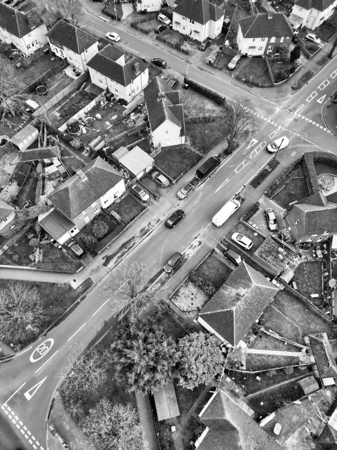 An aerial black-and-white photograph showing a residential street during a house removal process. The image captures multiple parked cars along the roadside, with some vehicles positioned on driveways and others on the street. Several houses with pitched roofs, varying in size and design, are visible, some with small gardens enclosed by fences or hedges. A large moving van is parked on the curb, with a loading area adjacent to a house, where furniture and cardboard boxes can be seen being moved or prepared for transport. Wrapping materials, such as plastic blankets and packing tape, are likely used to protect furniture during the home relocation. The scene indicates a busy moving day, with the loading process occurring carefully to facilitate efficient furniture transport. The environment appears clear and well-lit, emphasizing the logistics involved in domestic removals, consistent with services provided by Man with van Bedford Park.