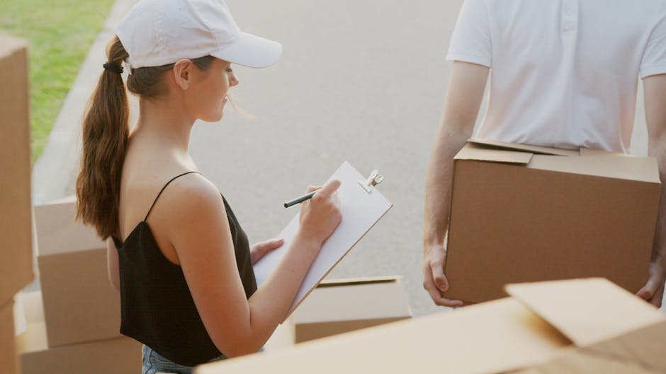 A woman with long brown hair tied in a ponytail, wearing a white cap and a black sleeveless top, is standing outdoors in front of a suburban street, holding a clipboard and pen, and appearing to take notes. She is positioned next to several cardboard boxes, some with packing materials, which are stacked on the pavement and inside a property doorway. A person in a white t-shirt, whose face is not visible, is carrying a medium-sized cardboard box, positioned to the right of the woman. The background includes a residential area with a paved road, greenery, and partial views of houses. This scene depicts a home relocation or moving process involving packing and furniture transport, which aligns with services offered by [COMPANY_NAME] in the house removals category, specifically related to loading, packing, and organizing a household move.