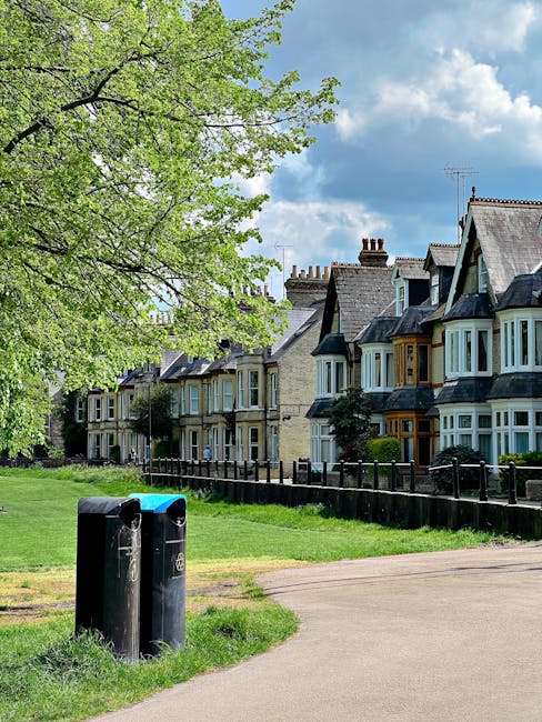 An outdoor scene on Devonshire Road showing a row of traditional terraced houses with bay windows, some with decorative woodwork, and chimneys, under a blue sky with scattered clouds. In the foreground, there are two black wheelie bins placed on a grassy verge near a paved footpath, with a black metal fence separating the lawn from the pavement. To the left, a large green tree with dense foliage extends over the scene, partially shading the area. This setting illustrates the typical residential environment where a house removal or relocation service from Man with van Bedford Park might operate, emphasizing the exterior environment, packing, loading, and furniture transport processes associated with moving homes in Bedford Park.