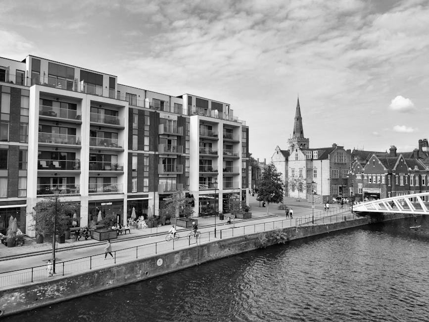 A black and white photograph showcases a modern residential building with multiple balconies and large windows situated along a riverside promenade in Bedford Park. The lower level of the building fea