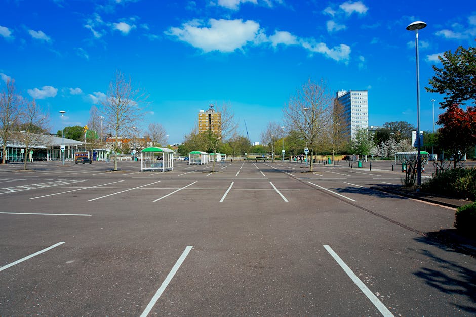 A spacious empty parking lot with clearly marked white parking bays, situated outdoors on a bright day with blue sky and scattered clouds. The lot is surrounded by leafless trees and some green bushes, with a few small structures or shelters visible in the background. Two tall street lamps are visible, one on the right and the other towards the left side of the image. In the distance, a few residential or office buildings of varying heights can be seen. The scene is well-lit, showing smooth tarmac pavement suited for vehicle loading and unloading, which is relevant for house removals and furniture transport logistics. Man with van Bedford Park uses such parking spaces for efficient home relocation and packing procedures, often assisting clients with vehicle access and loading processes in urban environments.