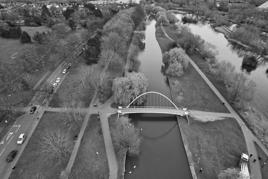 A black-and-white aerial photograph showing a small park area with winding pathways, trees, and a river flowing through it. A modern, curved pedestrian bridge with a white arch spans across the river, connecting two sides of the park. Several benches and lampposts line the pathways beneath the trees. On the left side of the image, a road runs parallel to the park with parked and moving cars. The park appears to be well-maintained, with neatly trimmed grass and leafless trees indicating a winter season. This scene captures a quiet urban environment, ideal for home relocation or moving logistics, with visible pathways suitable for loading and unloading furniture or boxes within the park area. The visibility of greenery, infrastructure, and transportation elements supports the context of professional removals services by Man with van Bedford Park, especially concerning transport planning for property moves in Bedford Park.