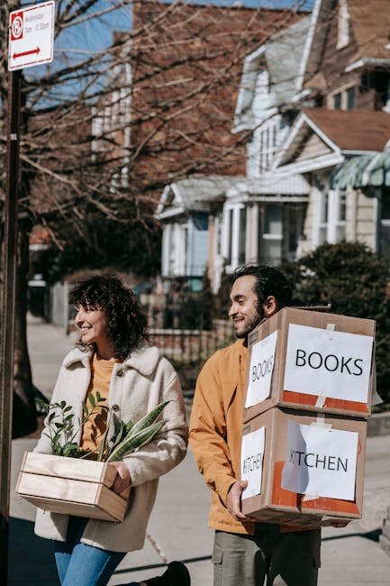 A man and woman smiling and carrying boxes inside a bright, modern living room with white walls, large arched window, and wooden flooring. The man is holding a small cardboard box labeled 'fragile' in