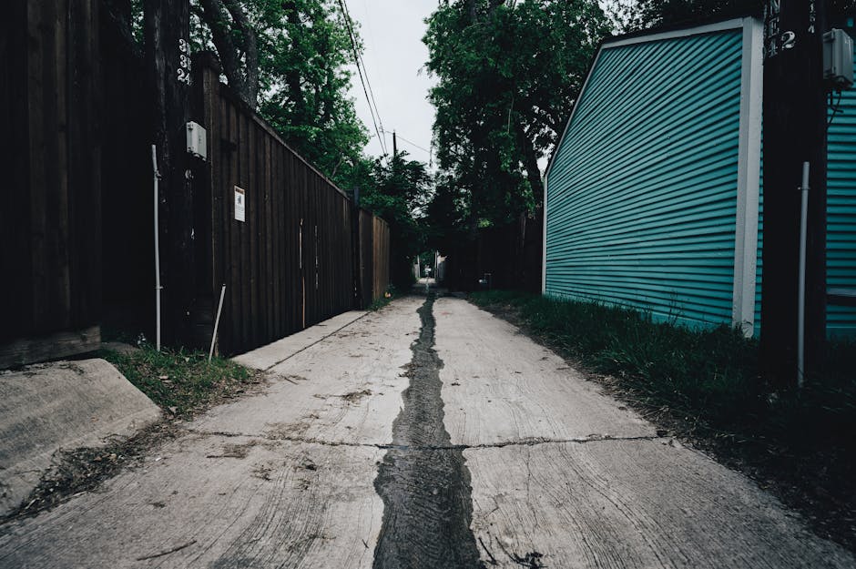 A narrow residential alleyway with a concrete surface shows visible cracks and a central drainage channel. On the left, there is a tall wooden fence with a small white sign and a utility box attached, while on the right, there is a blue corrugated metal building with a small patch of grass beside it. The alleyway is lined with lush green trees and bushes, with overhead power lines crossing above. The scene is illuminated by diffuse daylight, and no people or vehicles are visible. This setting represents the typical environment for home relocation and furniture transport, where careful planning is needed to avoid damage during the loading and unloading process. Man with van Bedford Park regularly manages such narrow street moves, supporting clients with efficient packing and transport services that ensure safe removal of household items within constrained urban environments.