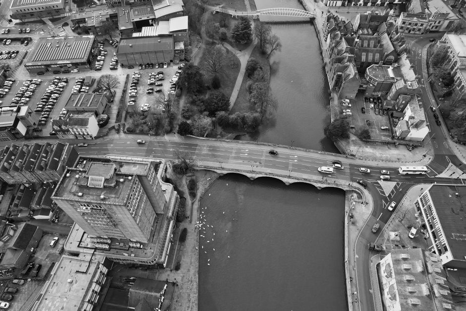 An aerial black-and-white photograph shows a cityscape featuring two body of water—one rectangular pond or small lake located in a park area with trees and a walking path along its edge, and the other a larger, irregularly shaped body of water beneath a road bridge. Adjacent to the water, there is a mix of modern and older commercial and residential buildings, some with flat roofs and others with sloped roofs. The road crossing the bridge has multiple lanes with several vehicles, including cars and vans, moving in both directions. Parking lots are visible nearby, with numerous parked cars. The surrounding environment includes paved walkways, small trees, and shrubbery, creating a typical urban landscape. The scene captures a moment during a home relocation or furniture transport process, with no visible moving activity, but highlighting the typical environment where [COMPANY_NAME] operates to assist with removals and packing between properties in Bedford Park. Natural light casts minimal shadows, indicating an overcast day.