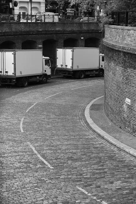 A black-and-white photograph shows a cobbled driveway with three white delivery vans parked parallel to a brick wall and a curved sidewalk. The vans are positioned near an archway beneath a raised area with a railing, overlooking a street with trees and buildings in the background. The driveway is marked with dashed white lines indicating parking spaces, and the scene appears to be in an urban residential or commercial area. The image captures a loading area suitable for home relocation or furniture transport, relevant to the services offered by Man with van Bedford Park. The vans are used for moving household items, and their proximity to the entrance suggests an ongoing or upcoming move, with potential loading or unloading activities, although no personnel or equipment are visible in this shot.